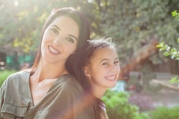 Young mom and daughter sitting back to back in park on sunny day and laughting