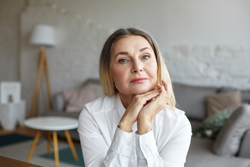 Head and shoulders of beautiful graceful senior woman with healthy wrinkled skin and loose thick hair having rest indoors, posing in modern interior, looking at camera with thoughtful face expression