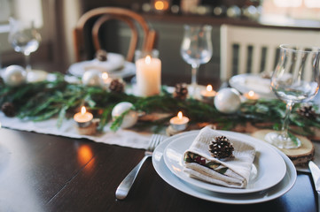 Festive Christmas and New Year table setting in scandinavian style with rustic handmade details in natural and white tones. Dining place decorated with pine cones, branches and candles