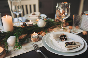 Festive Christmas and New Year table setting in scandinavian style with rustic handmade details in natural and white tones. Dining place decorated with pine cones, branches and candles