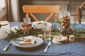 Festive Christmas and New Year table setting in scandinavian style with rustic handmade details in natural and white tones. Dining place decorated with pine cones, branches and candles