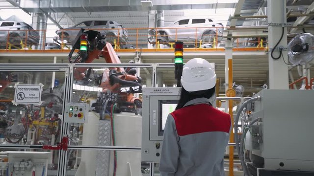 Automobile plant, modern production of cars, robot equipment, working woman adjusts production line machine control panel.
