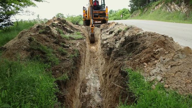 Excavator operating and digging soil for new electricity line. A backhoe tractor works on a construction site in open area green field.