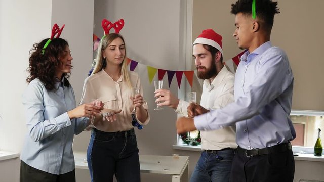 Multiracial group of happy office workers dancing during corporate New Year party wearing christmas hats and deer headband, holding glasses with sparkling wine and clinking
