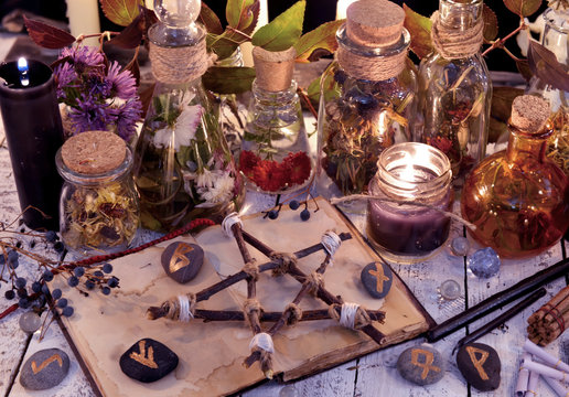 Wooden pentagram, open book, glass bottles, flowers, candles and runes on witch table