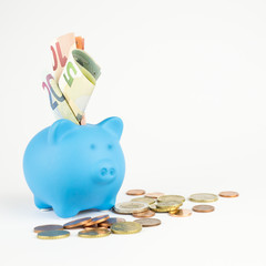 Ein blaues Sparschwein mit vielen Geldscheinen im Schlitz und Euromünzen daneben, quadratisch