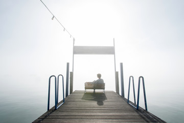 Obraz premium Frau sitzend auf kleiner Bank auf Holzsteg im Nebel hinaus blickend auf See