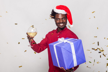 Around happiness afro man in red cap, flies gold metaphane, man holding champagne glass and gift box, looking at camera and toothy smiling.