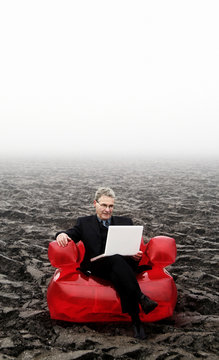 The Red Chair Business Serie: Senior Businessman In A Field On A Big Red Seat Working On His White Laptop