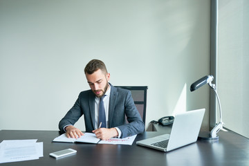 Waist-up portrait of confident young entrepreneur in classical suit taking necessary notes while preparing for important negotiations with business partners, interior of modern office on background
