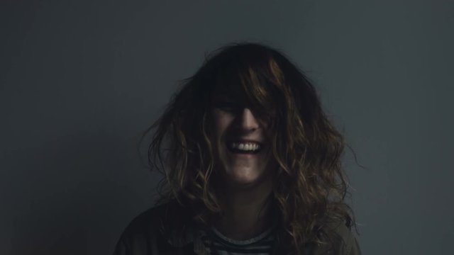 A woman standing behind a grey wall makes various happy expressions and gestures: thumbs up, hair playing, hair ruffling, and smiling. Close up.