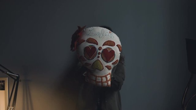 A skull shaped and patched pillow being held up by a person standing behind a grey wall. Medium shot.