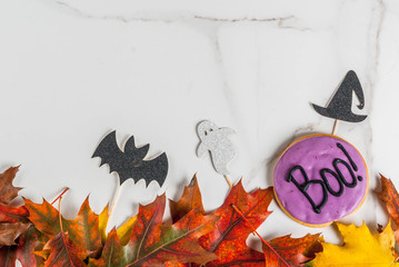 Festive background for Halloween, white marble table with gingerbread cookie with the inscription Boo!, holiday symbols (bat, witch hat, ghost) and autumn red yellow leaves, top view copy space frame
