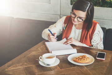 Young businesswoman in glasses and white sweater is sitting in cafe at table, working. Girl analyzes data, plans, considers income. Student learning online. On table cup of tea,smartphone. E-learning.