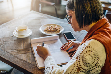 Side view. Young woman in eyeglasses is sitting in cafe at table,signing documents. Girl writes pen, fills out an application, questionnaire. Student is doing homework.On table cup of tea, smartphone.