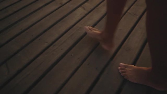 cropped picture of female tanned legs walking along wooden jetty barefoot during amazing sunset with colorful reflection of sun on water background in slow motion close up