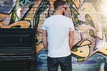 Summer day. Back view. Young bearded hipster man dressed in white t-shirt is stands against wall with graffiti. Mock up. Space for logo, text, image. Instagram filter, film effect.