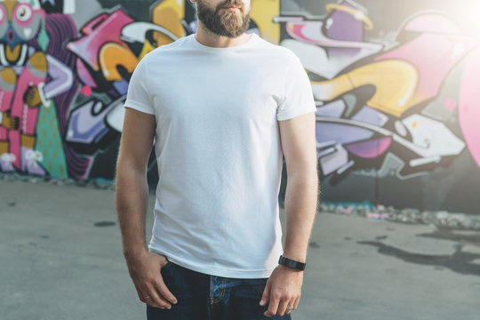 Summer day. Front view. Young bearded hipster man dressed in white t-shirt is stands against wall with graffiti. Mock up. Space for logo, text, image. Instagram filter, film effect, bokeh effect.