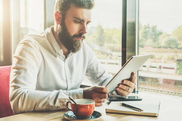 Young bearded businessman sits in cafe at table, holding tablet computer, working.On table cup of...