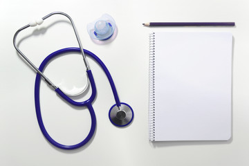 Stethoscope on a white table of the doctor's office, next to a pacifier, a note book and a pencil. Empty copy space for Editor's content. Top view.