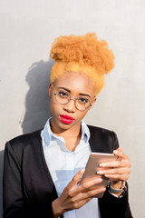 Fototapeta premium Lifestyle portrait of an african businesswoman in casual suit using smart phone on the gray wall background