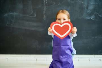 Portrait of adorable little girl holding red paper heart posing against blank blackboard in school, copy space