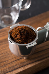 Pile of freshly roasted ground coffee in metal filter of the espresso machine on wooden surface ready to go to espresso machine with empty glass waiting