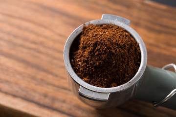 Pile of freshly roasted ground coffee in metal filter of the espresso machine on wooden surface ready to go to espresso machine with empty glass waiting