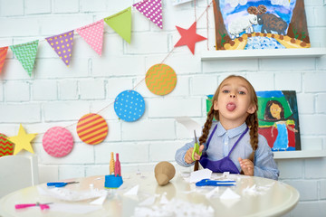 Portrait of pretty little girl looking at camera while making handmade Christmas decorations during art and craft class of pre-school and having fun sticking out tongue.