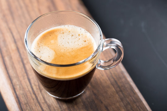 Glass of americano espresso fresh made dark black strong coffee on wooden board table, thick froth foam in cafe, morning refreshing hot drink caffeine, copy space for text