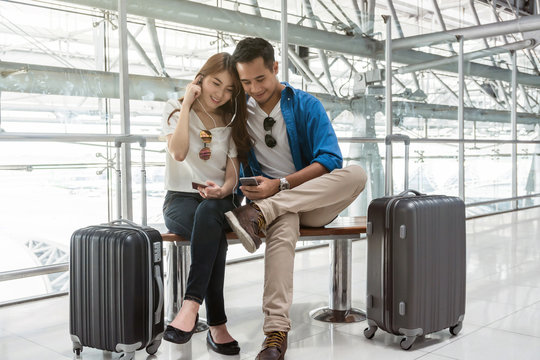 Asian couple traveler using smartphone and listening the song when waiting the airplane arrive with luggage at the airport. Lover travel and transportation with technology concept.