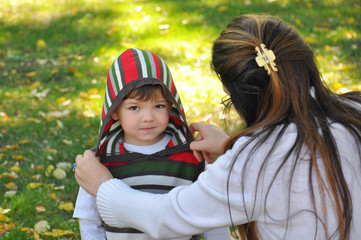 Mother get dressed her son in autumn park. Mom help her son with hat