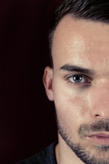 Obraz premium Portrait of beautiful young man with green eyes on dark black and red background. Half face portrait of man with beard and sharp lines of face. Attractive guy half face close up photo.