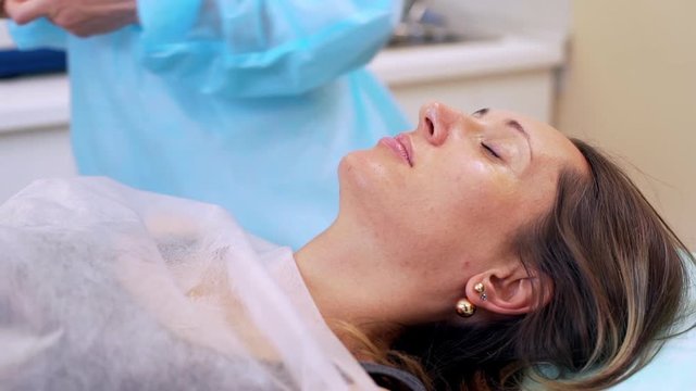 closeup picture of attractive woman lying on table with closed eyes and female arms putting gel around her eyes before botox reallife
