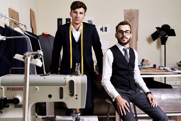 Portrait of two  handsome tailors posing in traditional atelier studio behind sewing machines , looking at camera