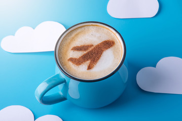 Cup of cappuccino with a picture of the plane on the foam. Blue sky background with paper clouds. Concept of travel