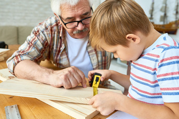 Portrait of nice senior man helping little boy build birdhouse, teaching him woodwork at table in...
