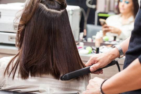 Stylist Using Flat Iron On Hair Of Female Client Sitting In Salon Chair