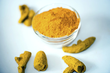 A glass bowl filled with bright yellow turmeric powder, surrounded by dried turmeric roots on a white surface. The image highlights the vibrant color and texture of this aromatic spice