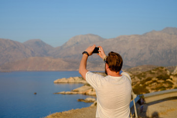 Naklejka na ściany i meble Young man makes selfie with stunning views of sea and mountains. View from the back.