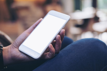 Mockup image of woman's hands holding white mobile phone with blank screen on thigh in cafe