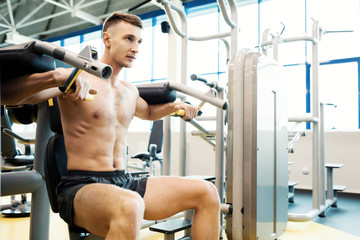 Fototapeta na wymiar Portrait of handsome young man with bare chest pumping arm muscles doing exercises on machines in gym