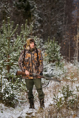 Male hunter with backpack, armed with a rifle, standing in a snowy winter forest.