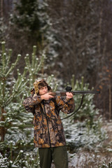 Fototapeta premium Male hunter in camouflage and with backpack, armed with a rifle, standing in a snowy winter forest.