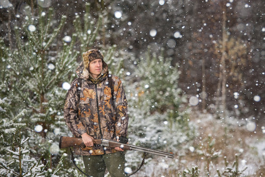 Male hunter with backpack, armed with a rifle, standing in a snowy winter forest. Man looks away. Snowfall in the forest.