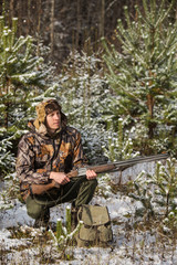 Fototapeta premium Male hunter in camouflage, armed with a rifle, sitting in a snowy winter forest. Man is charging a hunting rifle. Winter snow-covered forest.