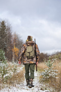 Male hunter in camouflage and with backpack, armed with a rifle, walks through the snowy winter forest.