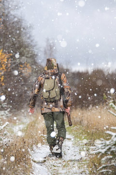 Male hunter in camouflage and with backpack, armed with a rifle, walks through the snowy winter forest.