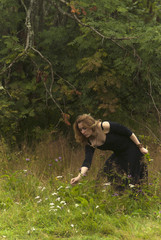 red-haired girl in a black dress collects herbs at the edge of the forest