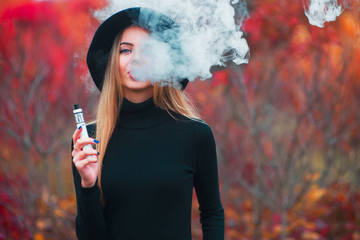 Young beautiful girl inblack hat vaping vape device with cloud of vape outdoors in autumn.